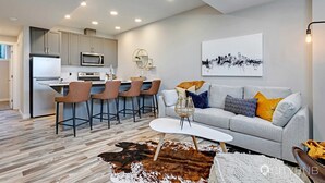 A modern kitchen with a dining area, a living room with a sofa and a cityscape painting, and a unique cowhide rug. - Blatchford - 2 Queen Beds - W/D to NAIT (Northwest)