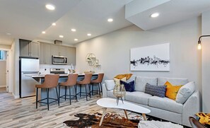 A modern kitchen with a dining area, a living room with a sofa and a cityscape painting, and a unique cowhide rug. - Blatchford - 2 Queen Beds - W/D to NAIT (Northwest)