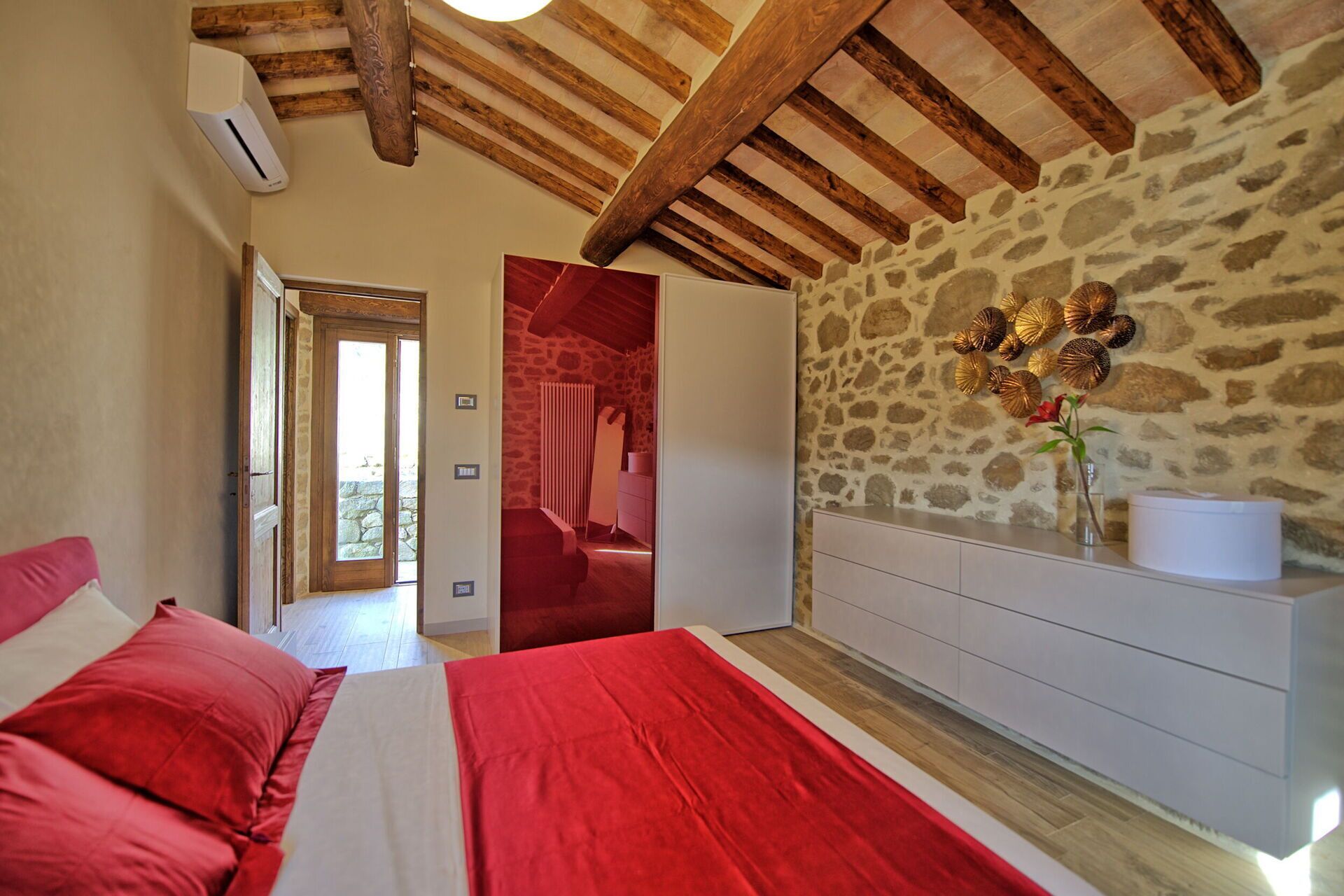A bedroom with a stone wall, wooden beams, a bed with red bedding, a dresser, and a vase with flowers.