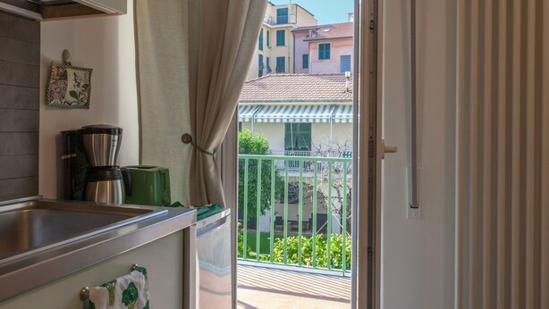 A kitchen with a sink, a coffee maker, and a green appliance. A curtain is partially drawn, revealing a balcony with a railing and a view of buildings.