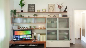 A living room with a flat-screen TV, shelves with decorative items, a cabinet with glass doors, and a door leading to another room.