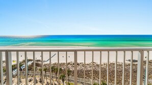 Vista a la playa desde un balcón con barandilla, playa arenosa y agua azul cristalina.