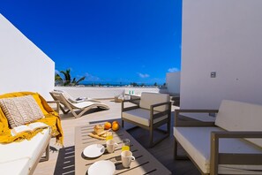 A sunlit outdoor seating area with lounge chairs, a table with breakfast items, and a view of the ocean.