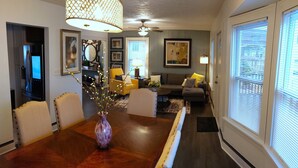 A dining area with a wooden table, chairs, a vase with flowers, a ceiling fan, and a window with blinds.