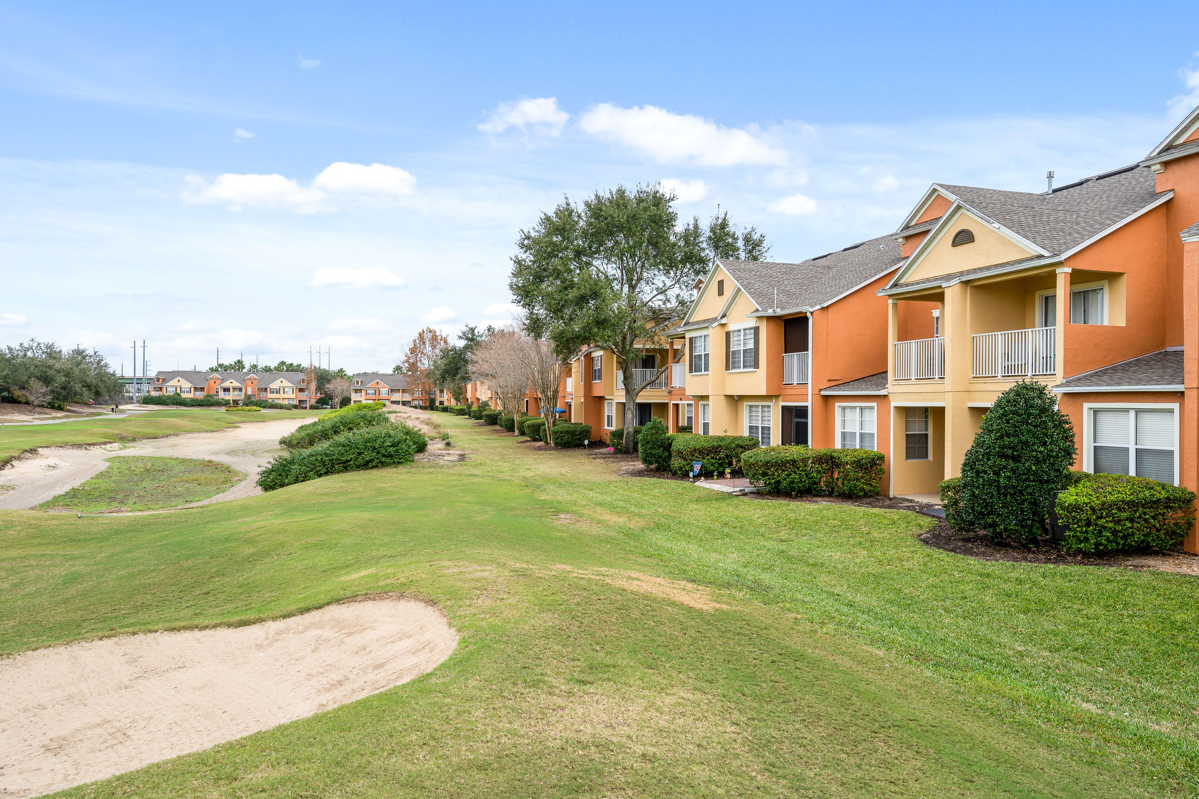 A row of multi-story residential buildings with balconies, a well-maintained lawn, and a clear sky.