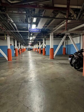 Un parcheggio multipiano con dissuasori arancioni, una segnaletica chiara che indica la direzione dell'ascensore e uno scooter parcheggiato a destra.