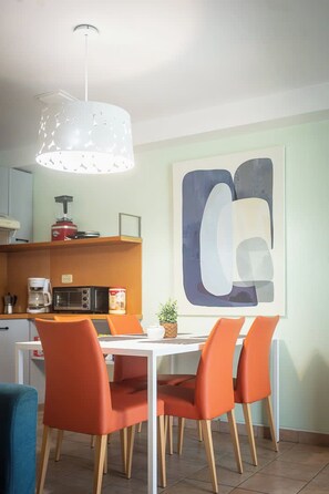 Zona pranzo con tavolo bianco, sedie arancioni, lampadario e un grande dipinto astratto appeso al muro.
