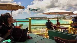 Un restaurante frente a la playa con mesas al aire libre, con vista al océano y un avión a lo lejos.