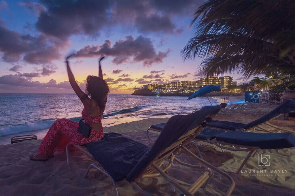 A beach scene with a person sitting on a lounge chair, a palm tree, and a building in the background.