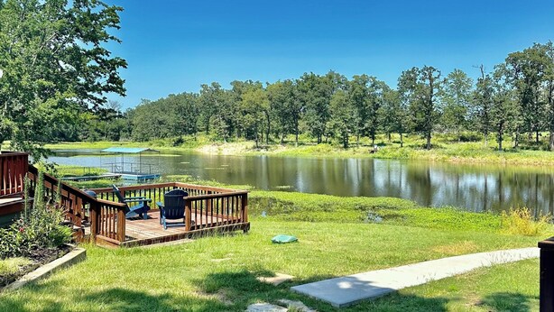 A deck with outdoor seating overlooking a lake, surrounded by greenery and trees. - SweetDove Stays-  Suite; w/ Stocked Lake, Secure Parking (Rains County)