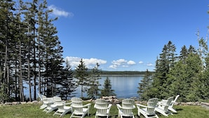 Vista del lago con sillas Adirondack blancas dispuestas en semicírculo frente al agua, rodeado de altos árboles y un cielo despejado.