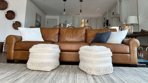 A living room with a brown leather sofa, white and blue cushions, and two white poufs with fringe.