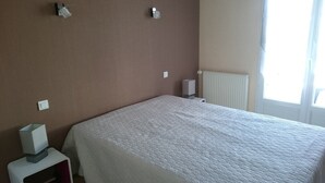 Una habitación de hotel con una cama grande, una mesita de noche con lámpara, un radiador y una ventana con cortinas.