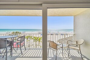 Un balcon avec des fauteuils en osier et une table, donnant sur une plage parsemée de parasols et animée par la présence de nombreuses personnes.