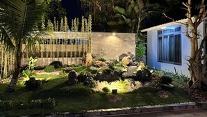A well-lit garden with a stone wall, a small building with a window, and various plants and rocks. - Prague Bungalow Phu Quoc (Phu Quoc)