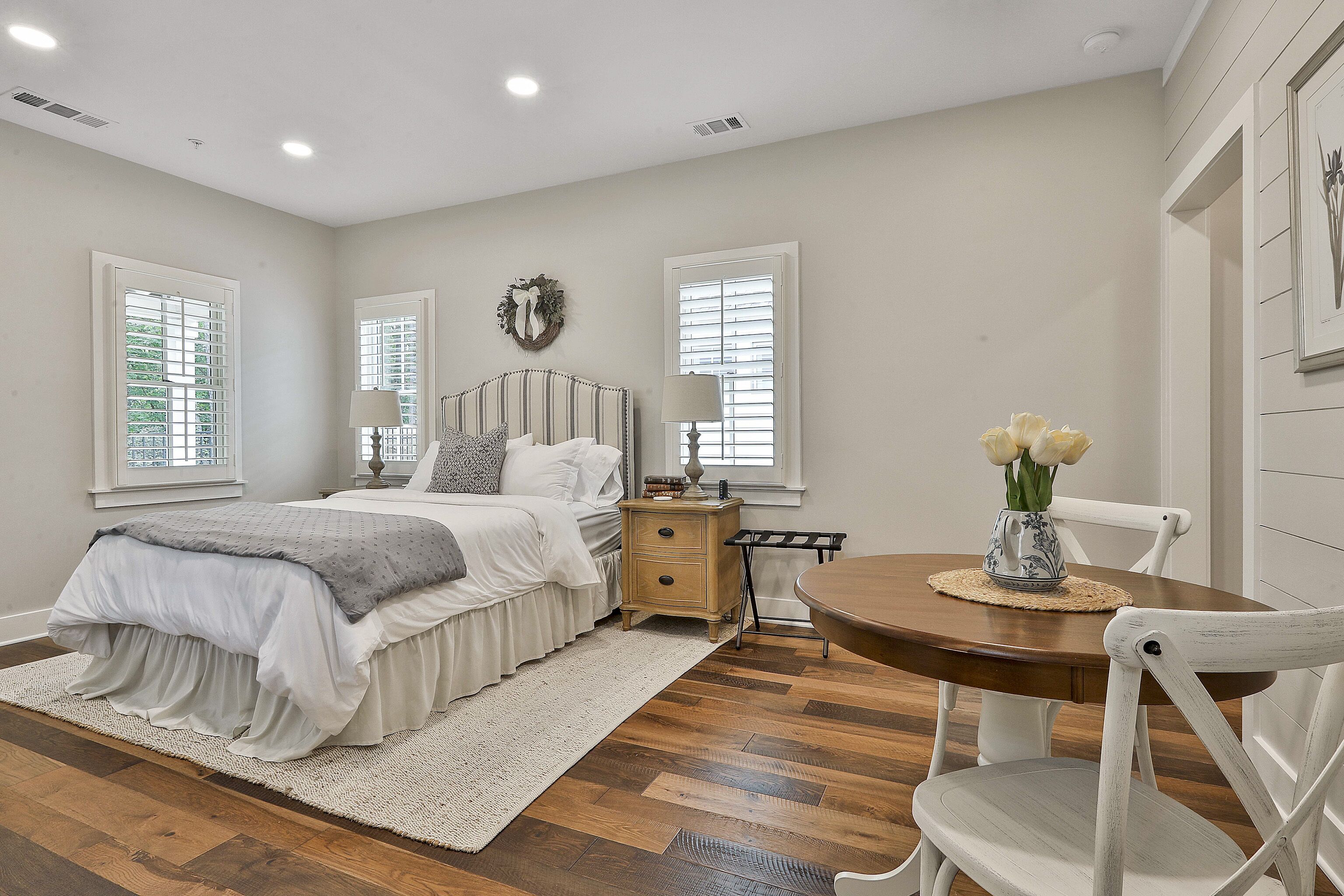 A bedroom with a bed, bedside tables, a nightstand, a chair, a table with a vase of flowers, and a window with blinds.