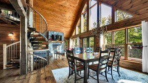 Een houten cabin interieur met een spiltrap, een stenen open haard, een eethoek met een tafel en stoelen, en grote ramen met uitzicht op de omgeving.