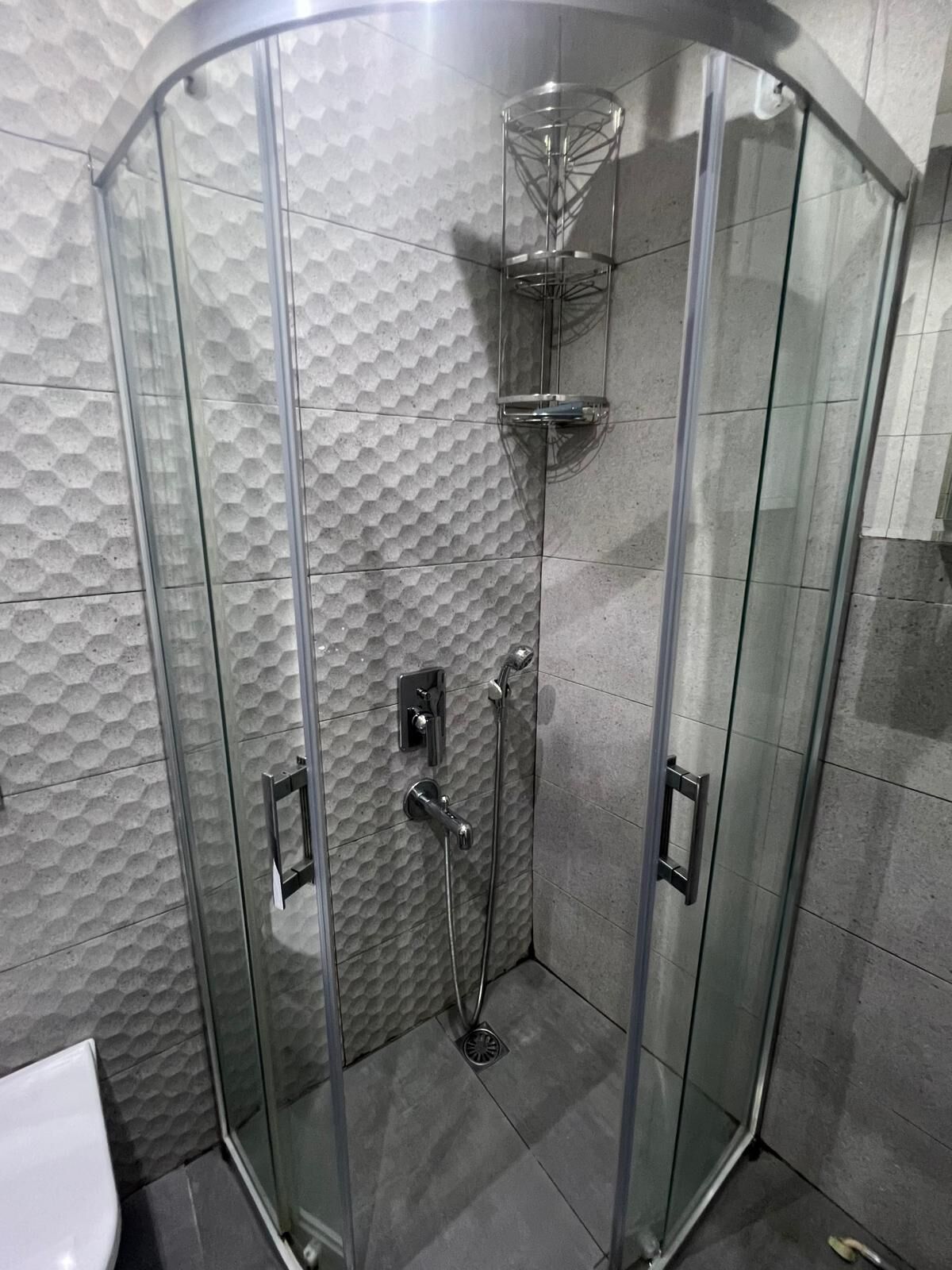 A glass-enclosed shower with a textured wall, a glass shelf, and a handheld showerhead.