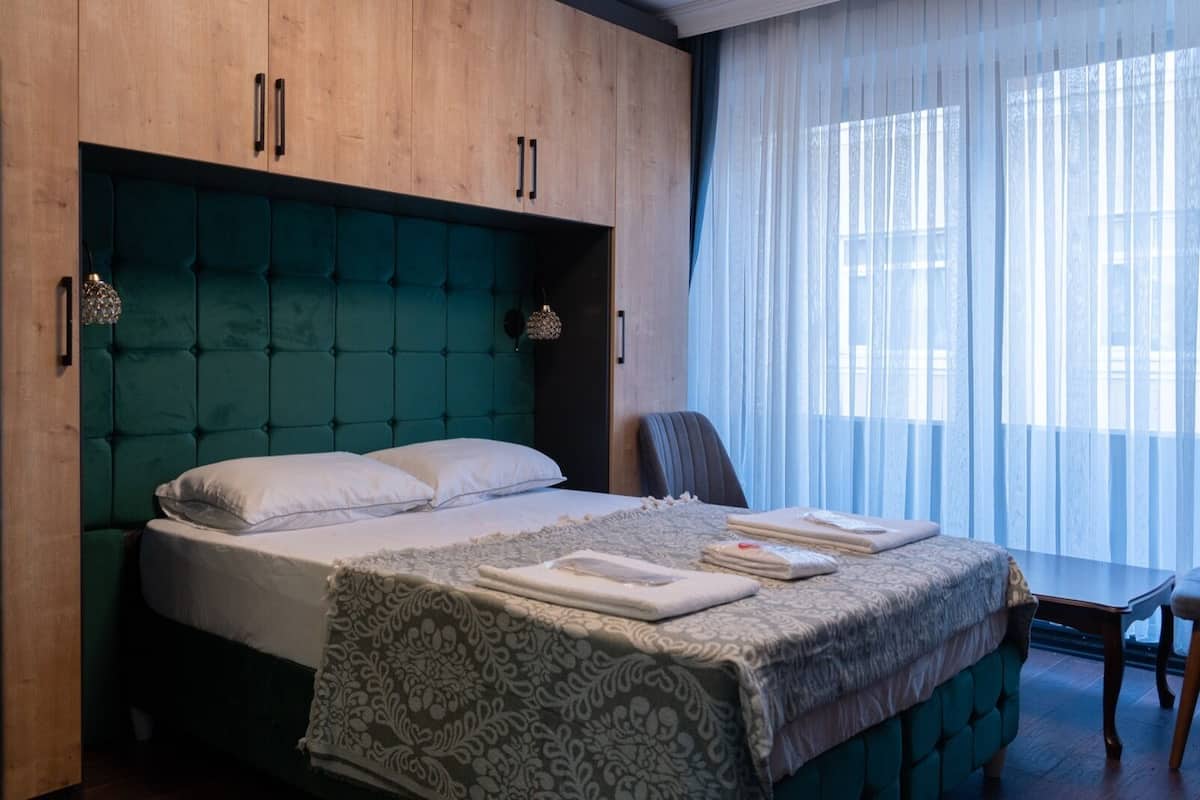 A hotel room with a large bed, a wooden wardrobe, a chair, and a window with sheer curtains.