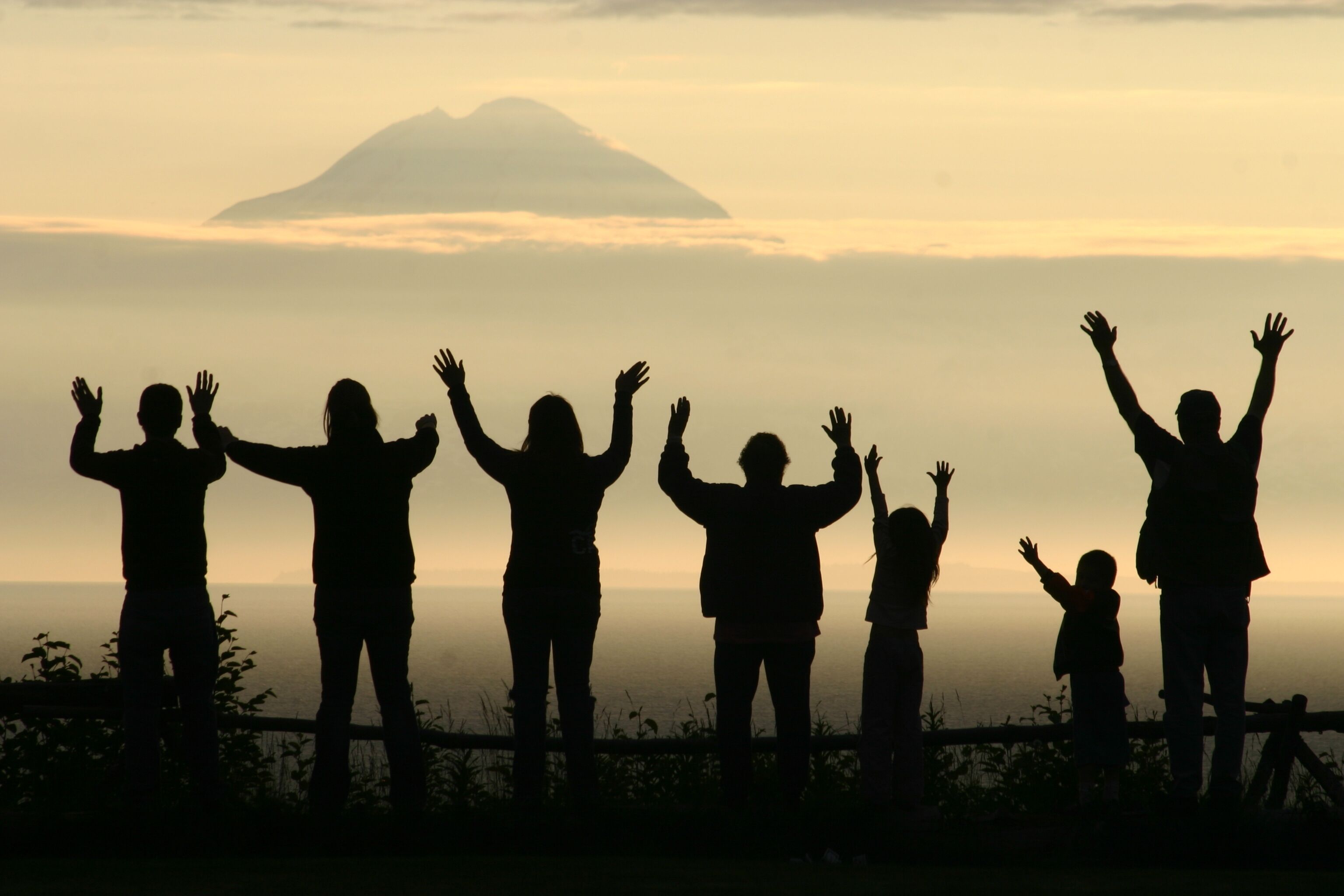 Silhouetten van mensen met geheven armen voor een zonsondergang en een berg op de achtergrond.
