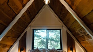 Un dormitorio con techo inclinado, cama con almohadas, ventana y luces fijadas a la pared.