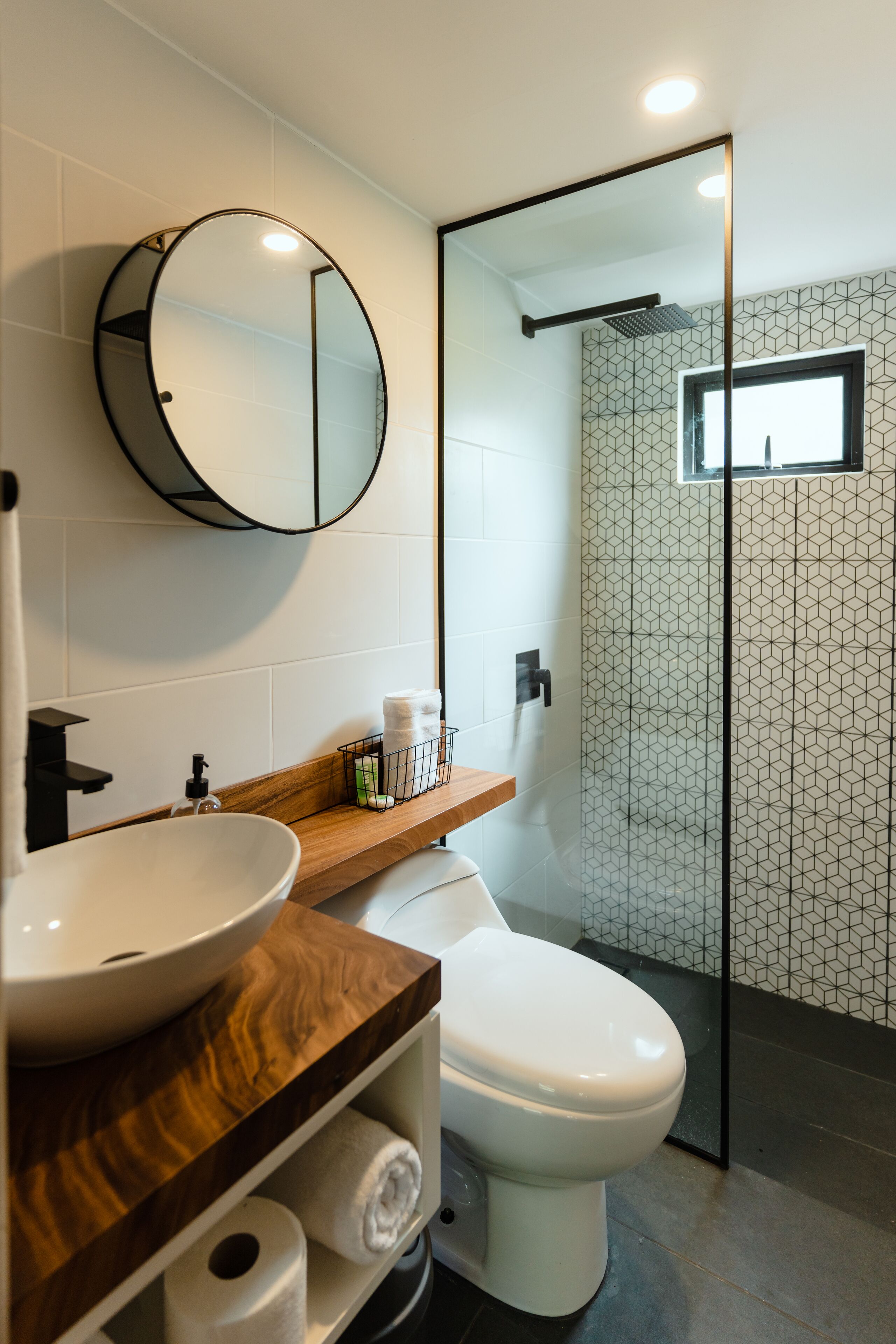 Un baño moderno con un espejo redondo, tocador de madera, inodoro blanco y ducha con mampara de vidrio.