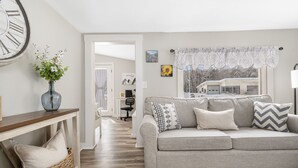 Una sala de estar con un sofá gris, una mesita de mimbre, un jarrón con flores, un reloj grande y una ventana con cortinas de encaje.