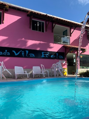 A pink building with a sign reading "Vila Bela" and a pool area with white chairs. - Pousada Vila Bela (Ipojuca)