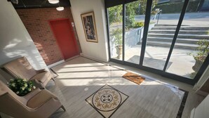 Une salle d’attente avec une porte rouge, un tableau encadré au mur et une vue sur l’extérieur grâce à de grandes fenêtres.