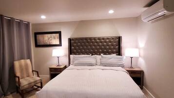 A hotel room with a large bed, two bedside tables with lamps, a chair, a framed picture on the wall, and an air conditioning unit.
