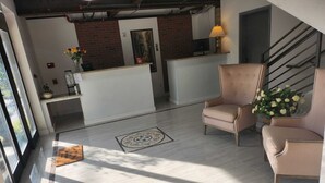 Un espace d’accueil avec un comptoir blanc, deux chaises beiges, une petite plante en pot et un escalier en arrière-plan.