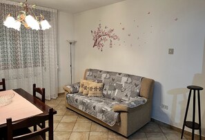 A living room with a sofa, a dining table, chairs, a chandelier, and a decorative wall mural.
