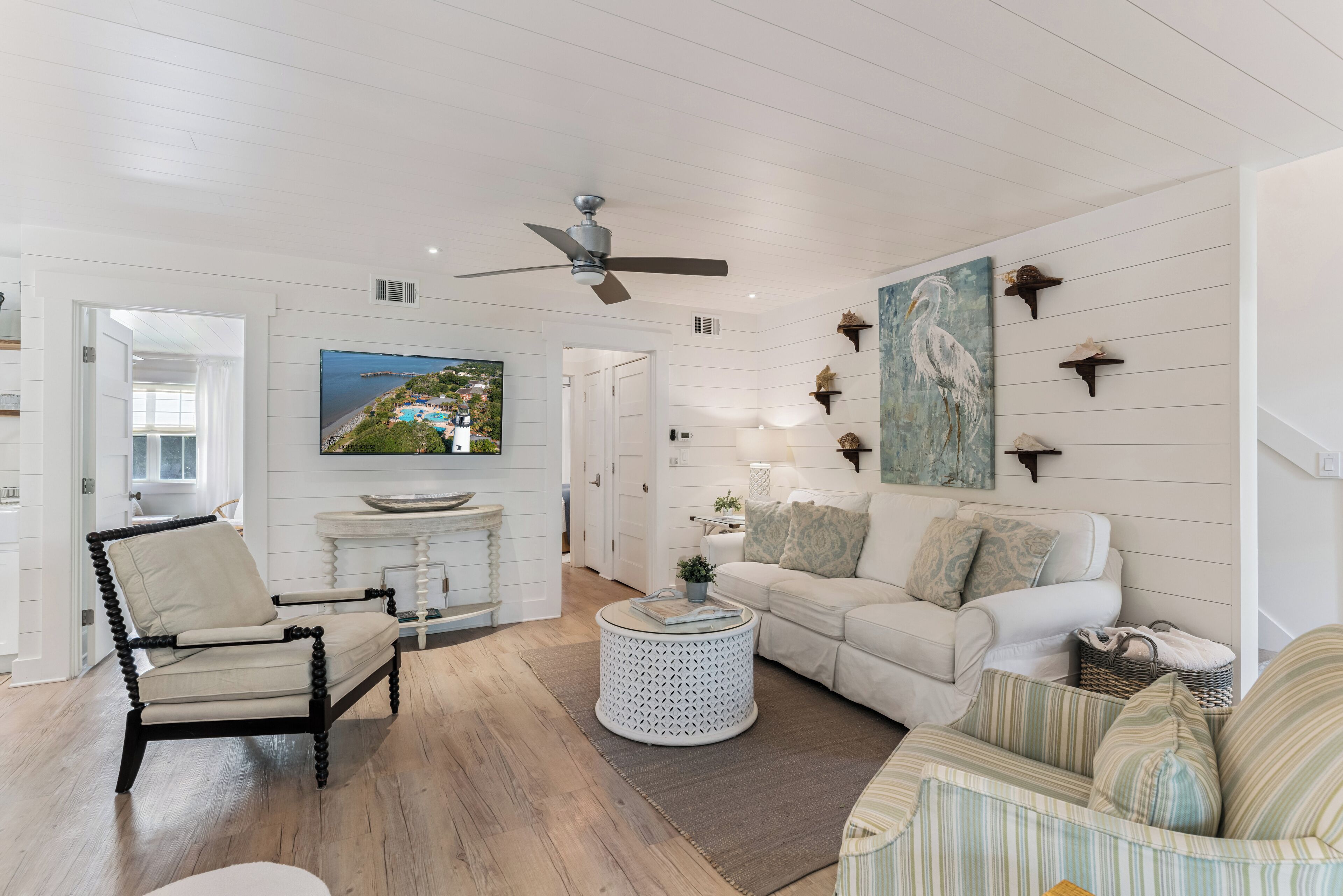 A spacious living room with a white sofa, a round coffee table, and a comfortable armchair. A television is mounted on the wall, and there are decorative wall shelves.