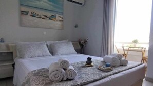 Habitación de hotel con una cama, un cuadro de barcos en la pared, una mesita con una planta y vista al mar por la ventana.