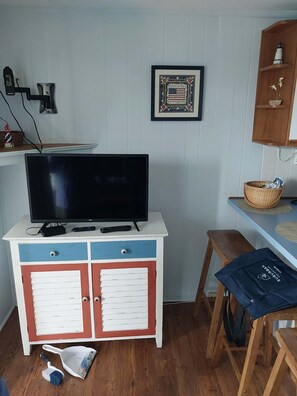A room with a TV on a white and blue cabinet, a framed picture on the wall, a wooden shelf with a bottle, and a chair with a bag. - Cape Cod Oceanside Condo across from beach! Combine World Cup + Beach Holiday! (Dennis)