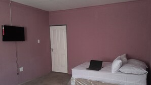 Une chambre à coucher avec un lit blanc, un ordinateur portable, un téléviseur fixé au mur et une porte fermée.