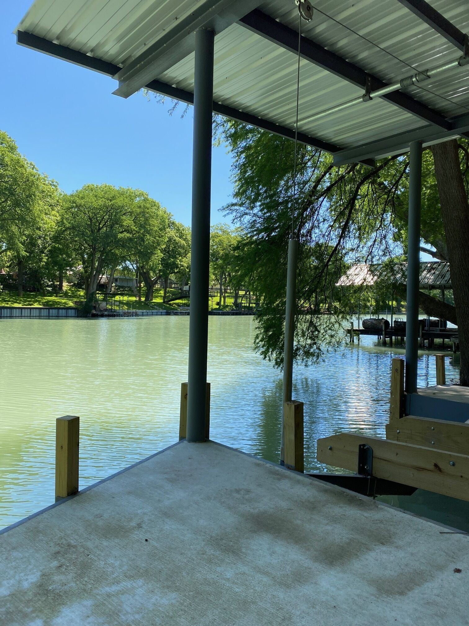 A covered outdoor area with a concrete floor, metal beams, and a view of a calm body of water with trees in the background.