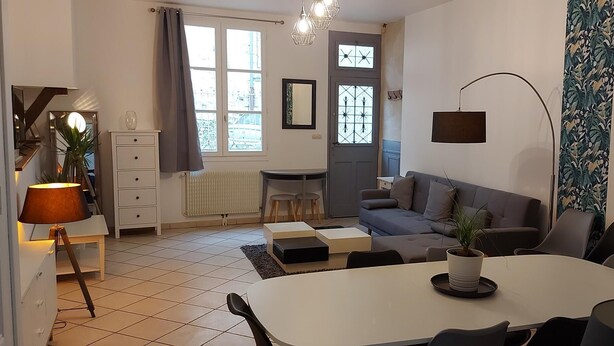 A modern living room with a white dining table, grey sofa, and a small side table. There is a potted plant on the dining table and a lamp on a side table.