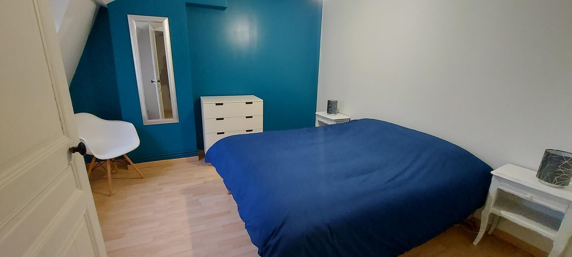 Una camera da letto con una coperta blu, una sedia bianca, un comodino e una finestra con telaio bianco.