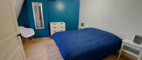 Una camera da letto con una coperta blu, una sedia bianca, un comodino e una finestra con telaio bianco.