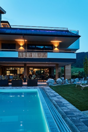 A modern two-story house with a swimming pool, outdoor seating, and a view of the surrounding landscape.