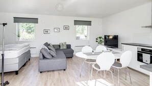 Un soggiorno moderno e luminoso con un divano grigio, un tavolo da pranzo bianco e sedie. Si vede anche una televisione e un angolo cottura.