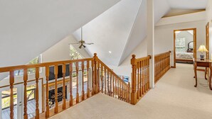 Une chambre spacieuse aménagée en mezzanine, avec un plafond haut, un escalier en bois et une grande fenêtre donnant sur l’extérieur.