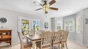 Un coin repas avec une table et des chaises en bois, un ventilateur de plafond et une vue sur l’extérieur, visible à travers la porte.