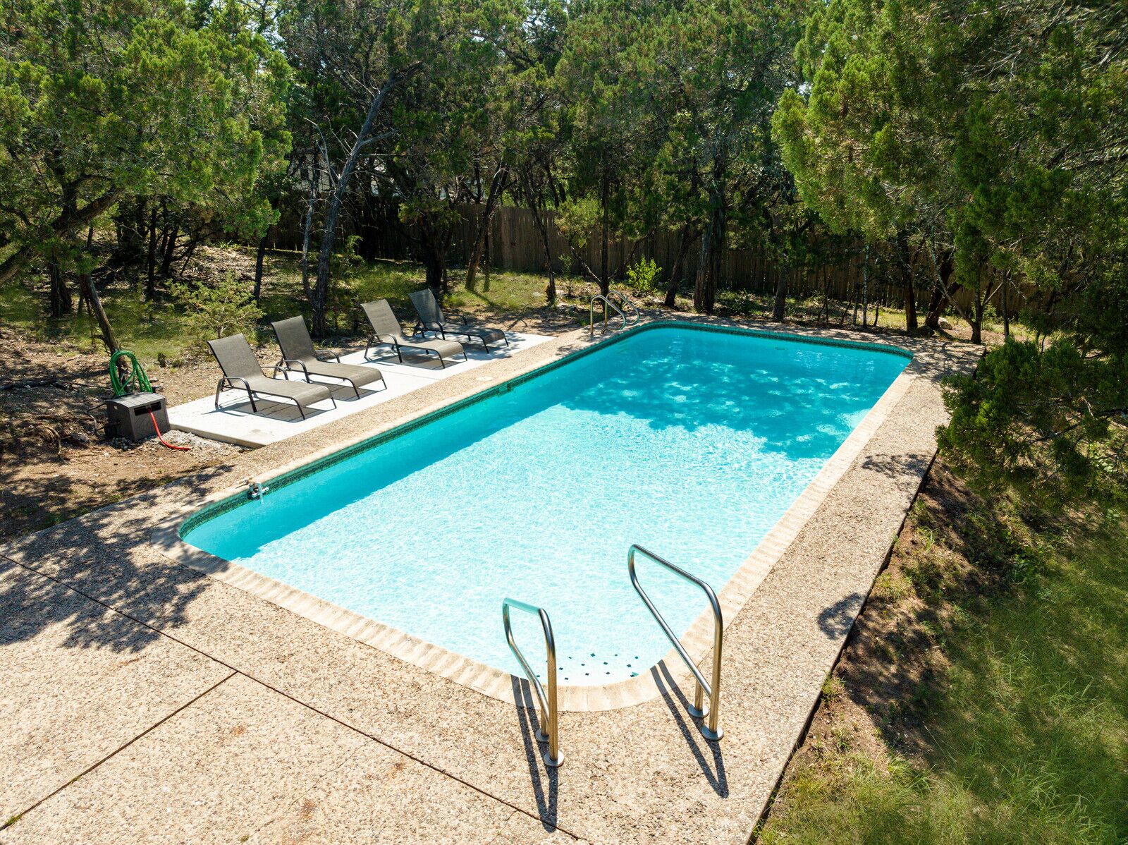 Ein rechteckiger Swimmingpool mit Fliesen umgeben, von Bäumen gesäumt und mit Liegestühlen und einer Leiter ausgestattet.