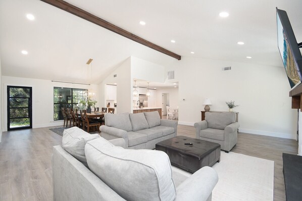A spacious living room with a large gray sectional sofa, a coffee table, and a TV mounted on the wall. There is a dining area with a table and chairs, and a kitchen in the background.