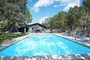 A rectangular swimming pool with clear blue water, surrounded by trees and a building with a dark roof.