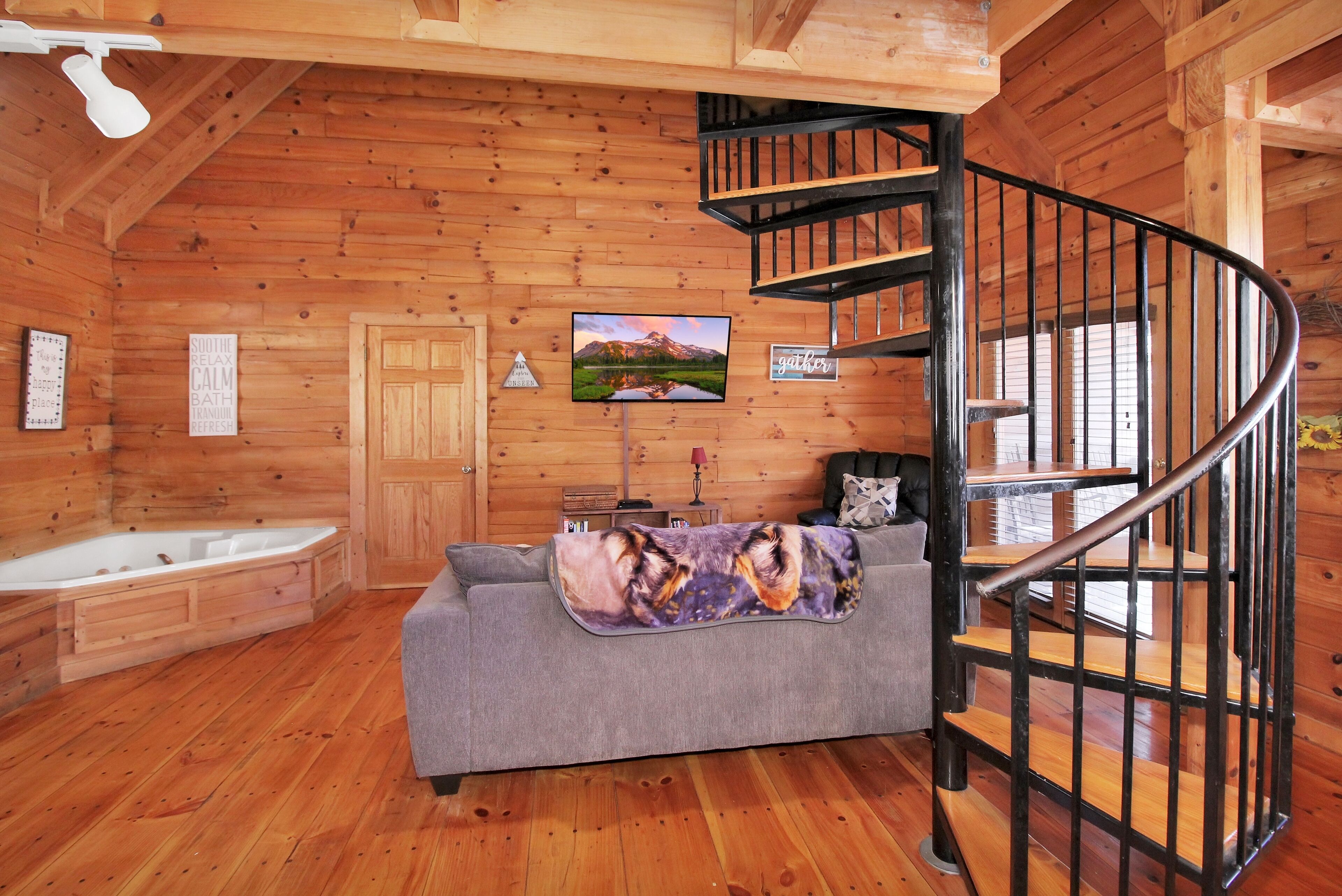 A wooden cabin interior with a gray sofa, a wooden staircase, a bathtub, and a framed picture on the wall.