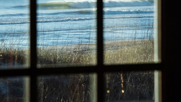 Blick durch ein Fenster mit Gittermuster auf einen Strand mit Wellen und einem klaren Himmel.