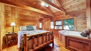 A wooden cabin room with a bed, a nightstand, a lamp, a window, and a bathroom with a bathtub.